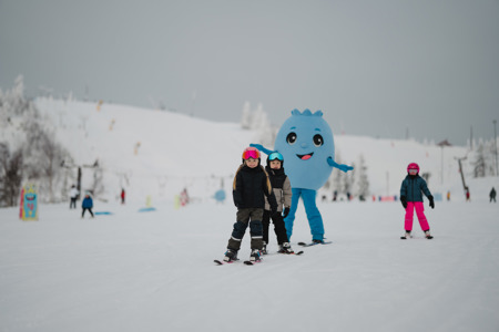 Barn åker skidor i en skidbacke tillsammans med en blå maskot i bakgrunden.
